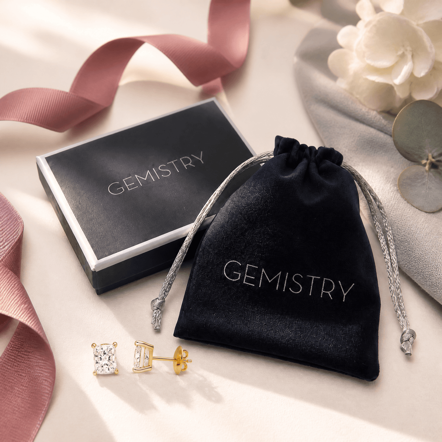 A pair of gold stud earrings with clear gemstones is displayed next to a black velvet drawstring pouch and a black presentation box, both printed with the brand name 'GEMISTRY'. The scene is styled with a dusty pink ribbon, a white flower, and a gray decorative disc on a light beige background.
