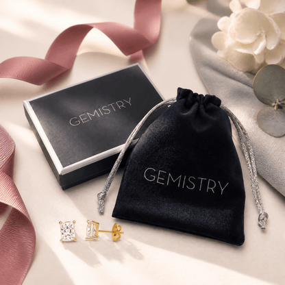 A pair of gold stud earrings with clear gemstones is displayed next to a black velvet drawstring pouch and a black presentation box, both printed with the brand name 'GEMISTRY'. The scene is styled with a dusty pink ribbon, a white flower, and a gray decorative disc on a light beige background.